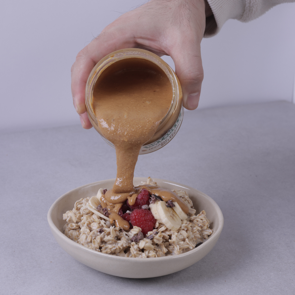 A hand pouring peanut butter from a glass jar onto a bowl of oatmeal topped with sliced bananas, berries, and chocolate chips on a light gray background.